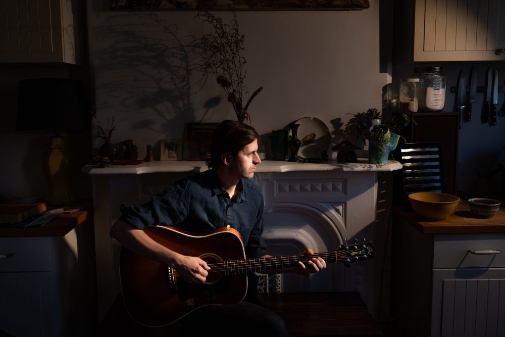 Man sits in a darkened room playing guitar and looking towards light