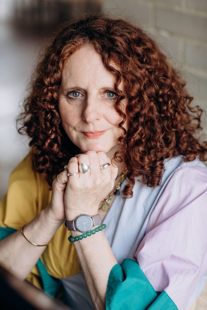 Photo of author Maggie O'Farrell. A woman with curly red hair rests her chin on her hands and looks directly at the camera||