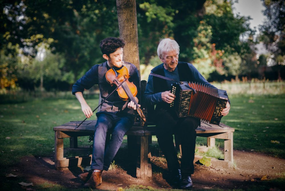 A father and son sit in a park playing fiddle and accordion together