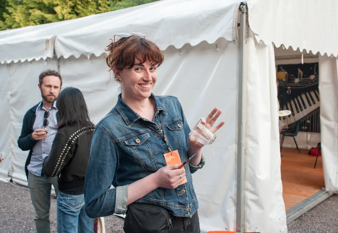 Woman holding a drink and smiling at an outdoor event beside a marquee tent.