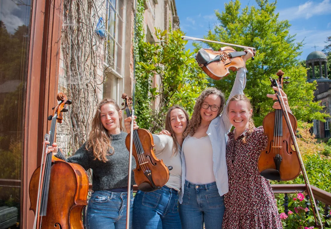 Four musicians holding string instruments outdoors in a garden setting.