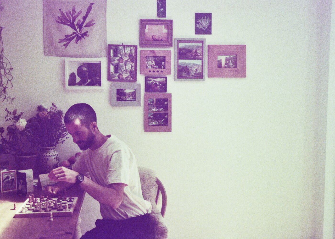 Grainy photo of a a man playing chess in front of a wall of pictures