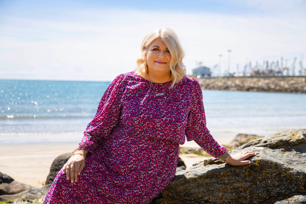 Photo of author Carmel Harrington in a purple patterned dress. She is sitting on a large rock at the beach with the blue sea and sky behind her